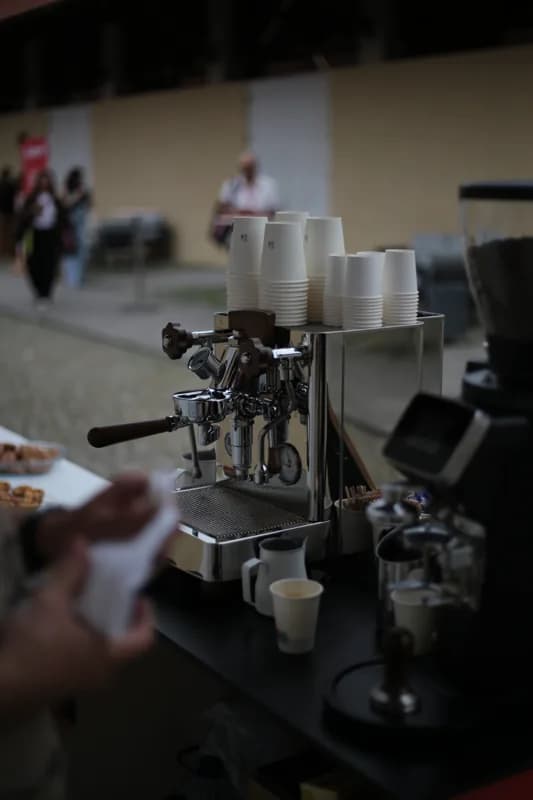 Máquina de café e copos em primeiro plano, com convidados e mesa de apoio desfocados ao fundo.